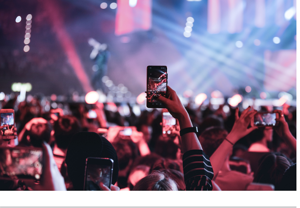 The picture shows a concert. Many in the audience are holding up their mobile phones and filming or photographing what is happening on stage.